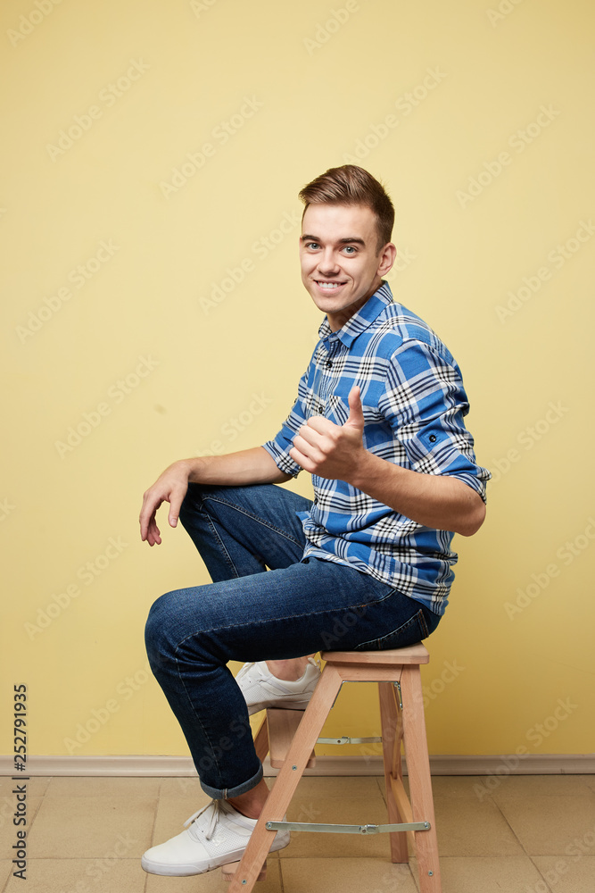 Cheerful guy dressed in a plaid shirt and jeans is sitting and giving thumbs up on a stool on a yellow background in the studio Cheerful guy dressed in a plaid shirt and jeans is sitting and giving thumbs up on a stool on a yellow background in the studio