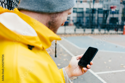Young man in yellow raincoat standing on the street at playground with mobile phone in hand 