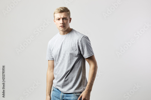 Serious blond guy dressed in a white t-shirt stands on the white background in the studio