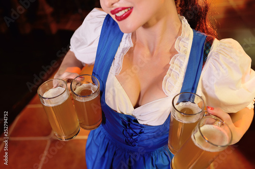 an attractive young girl in Bavarian clothes holds a lot of mugs with beer on the pub background during the celebration of OKtobfest