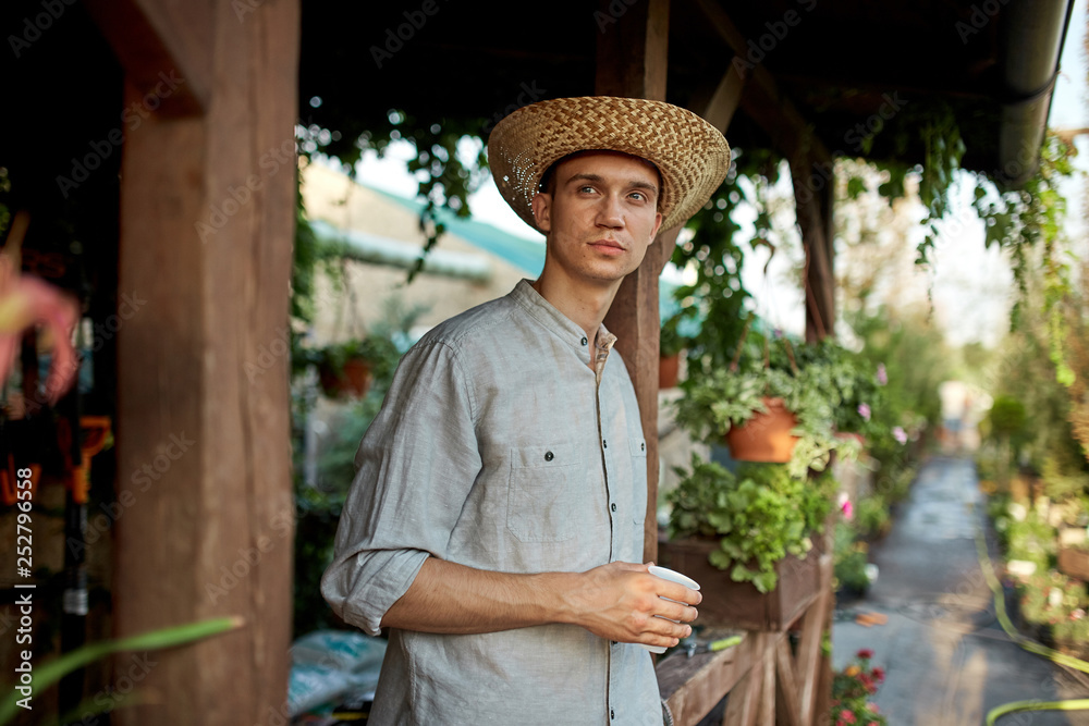 Guy gardener in a straw hat is standing with plastic glass in his hand next to a wooden veranda in the wonderful nursery-garden on a sunny day.