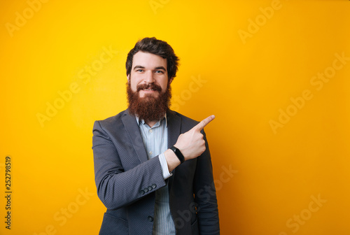 Image of young handsome businessman with beard pointing away with finger over yellow background