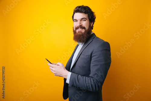 Portrait of a smiling young business man using smartphone and looking at camera isolated on a yellow background