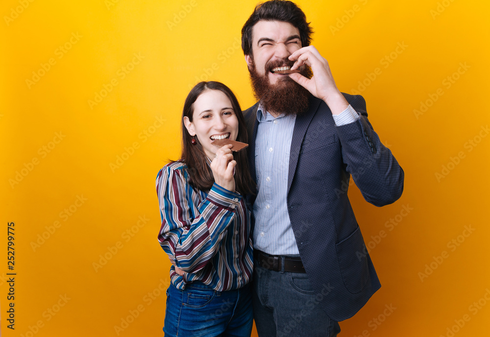 Young couple man and woman eating chocolate on isolated yellow background