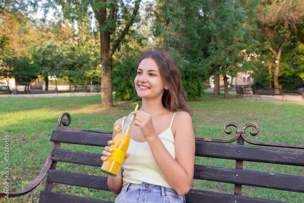 Fototapeta premium Young pretty girl is having rest on the bench in the park and drinking fresh juice from a bottle through the straw