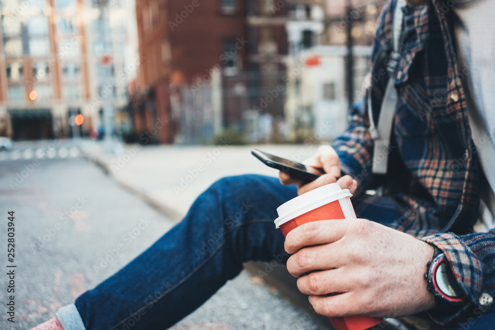 Man sitting on the street resting with cup of hot coffee and using mobile phone. Close-up