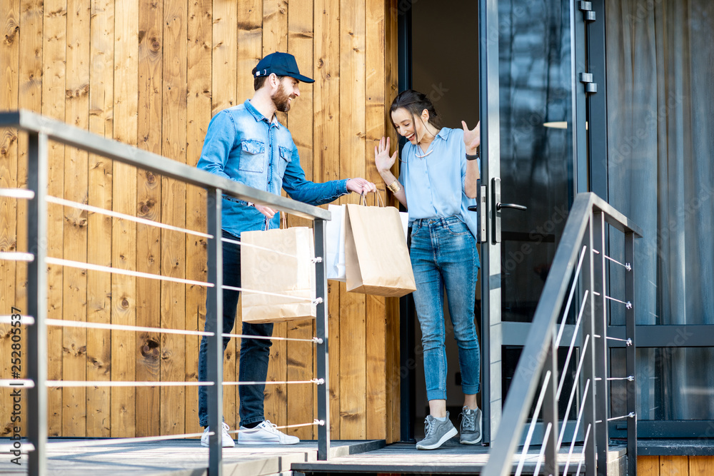 Delivery man bringing some goods packaged in paper bags for a young woman client to home. Buying clothes online and delivery concept