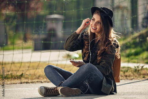 Smiling woman adjusting earphones while listening music on smart phone outdoors.