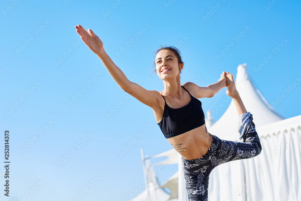 Portrait of a young slim athletic sports woman makes yoga exercises and stretching on the pier at sunny summer morning. Athletic lady doing Lord of Dance pose against blue sky.Natarajasana Yoga Pose Portrait of a young slim athletic sports woman makes yoga exercises and stretching on the pier at sunny summer morning. Athletic lady doing Lord of Dance pose against blue sky.Natarajasana Yoga Pose