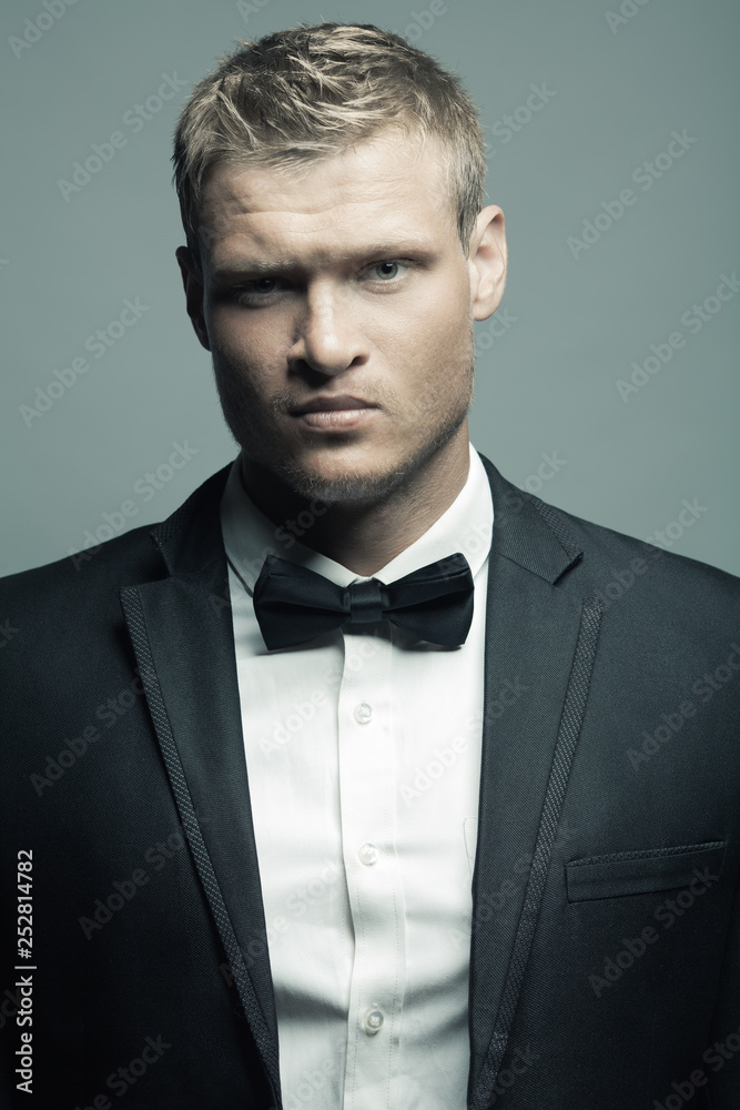 Male fashion, beauty concept. Portrait of brutal young man with short wet blond hair wearing black suit, posing over gray background. Classic style. Studio shot Male fashion, beauty concept. Portrait of brutal young man with short wet blond hair wearing black suit, posing over gray background. Classic style. Studio shot