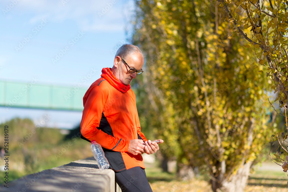 Side view of a senior runner man leaning on fence while testing exercise in a mobile phone outdoors in a sunny day Side view of a senior runner man leaning on fence while testing exercise in a mobile phone outdoors in a sunny day