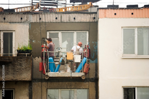 Two workers are plastering the facade of a building without observing safety techniques in a cradle on the upper floors of a high-rise building. The laborers are working without construction hardhat. © SVIATLANA