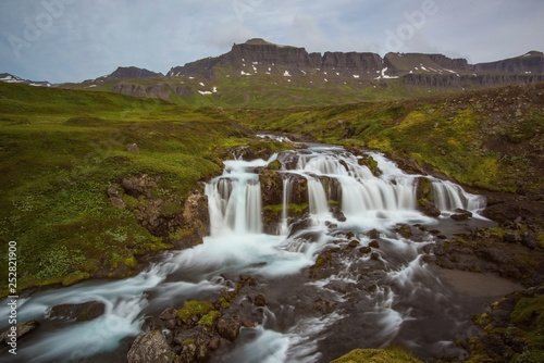 The Dyrfjollfoss Waterfall with golden clouds in the sky. The flowing water is captured by a long exposure. Amazing blue color of water from the glacier. Natural and colorful environment...