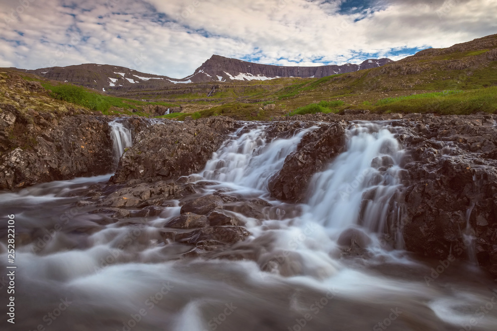 The Noname Waterfall with golden clouds in the sky. The flowing water is captured by a long exposure. Amazing blue color of water from the glacier. Natural and colorful environment...