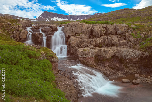 The Noname Waterfall with golden clouds in the sky. The flowing water is captured by a long exposure. Amazing blue color of water from the glacier. Natural and colorful environment...