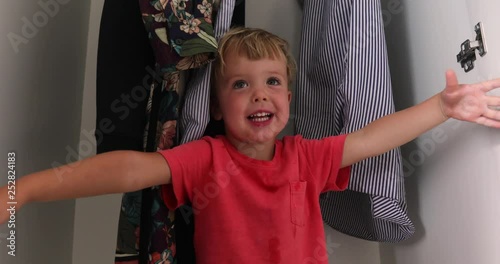 Side view of happy little boy playing in white wardrobe with shelves having fun at home