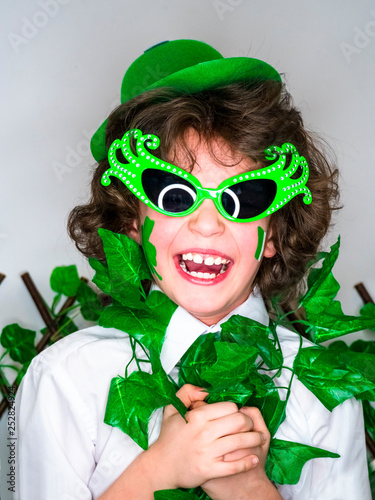 Child Celebrating St. Patrick's Day Showing his Make-up. A small, curly smiling boy in green carnival accessories looking at the camera. green background