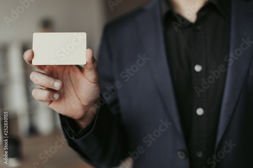 Close up of man in black suit holding white business card in hand. Blurred background. Cut view
