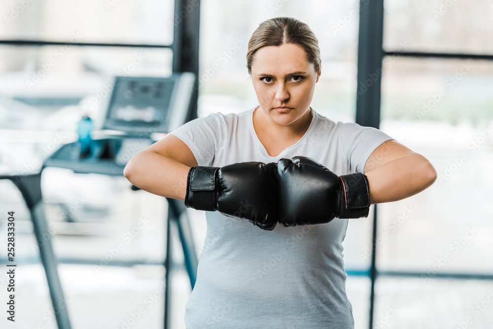 serious plus size woman wearing boxing gloves looking at camera in gym
