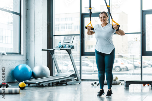 cheerful overweight woman training arms with suspension straps in gym