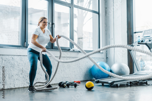 cheerful overweight woman training with battle ropes in gym photo