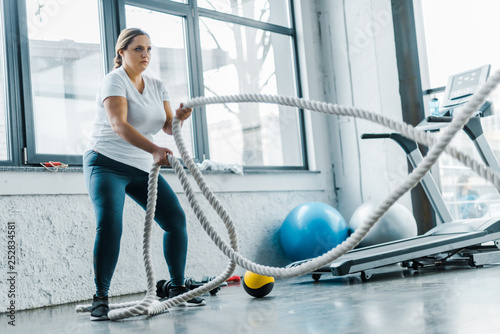 focused overweight woman training with battle ropes in gym