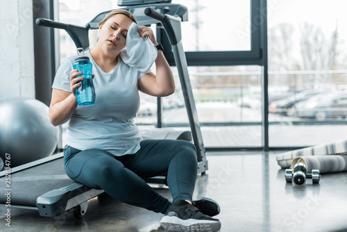 Fototapeta Naklejka Na Ścianę i Meble -  tired plus size woman wiping sweat with towel while sitting on treadmill and holding bottle with water