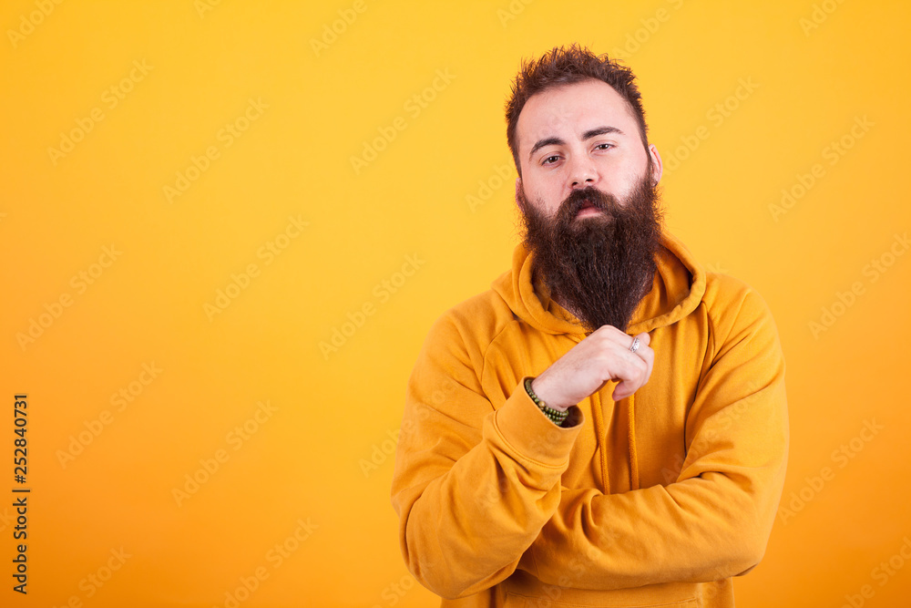 Stylish beaded hipster looking serios at the camera and playing with his beard over yellow background Stylish beaded hipster looking serios at the camera and playing with his beard over yellow background