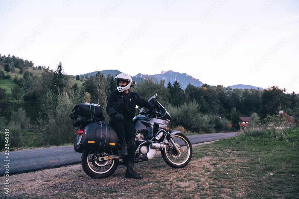 Rider Man and off tourism adventure motorcycles with side bags and equipment for long road trip, travel touring concept, Ceahlau, Romania, mountains on background, sunset evening