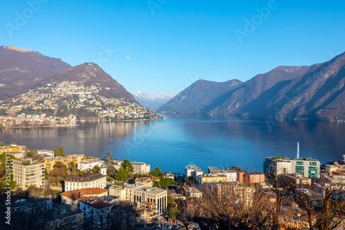 View over City and Alpine Lake Lugano with Mountain and the Moon in Switzerland.