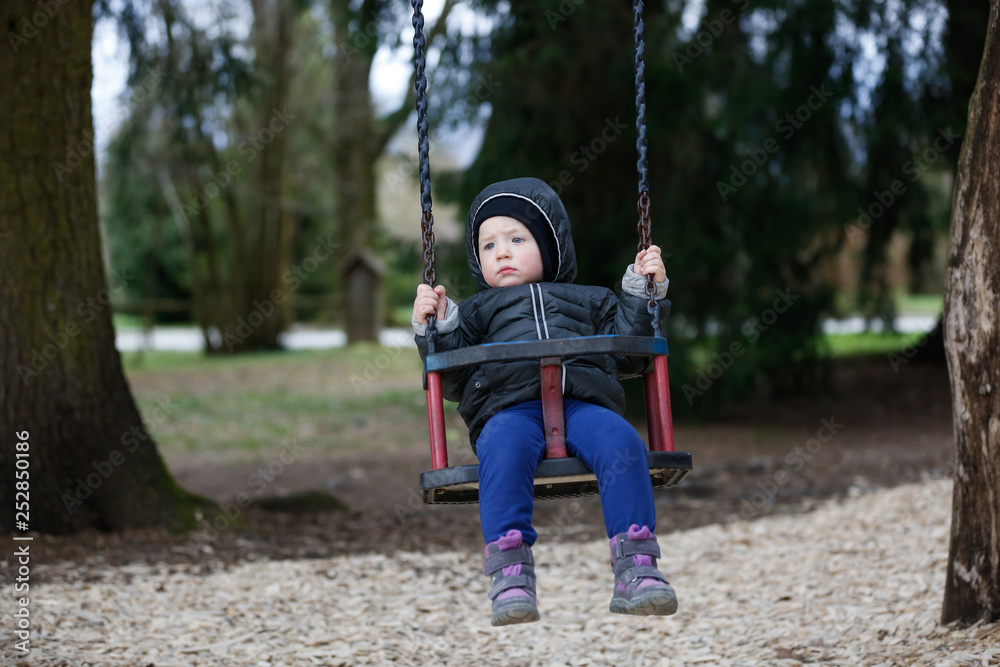 Sad, worried little boy swinging by himself, left alone unattended, looking for parents.