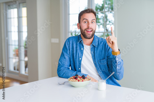 Handsome man eating cereals for breakfast at home pointing finger up with successful idea. Exited and happy. Number one.