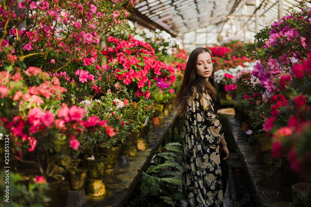 Young beautiful caucasian woman in glass greenhouse among colorful azalea flowers. Art portrait of a girl wearing a hat and romantic boho clothes. Gardening, plant growing, art therapy concept.