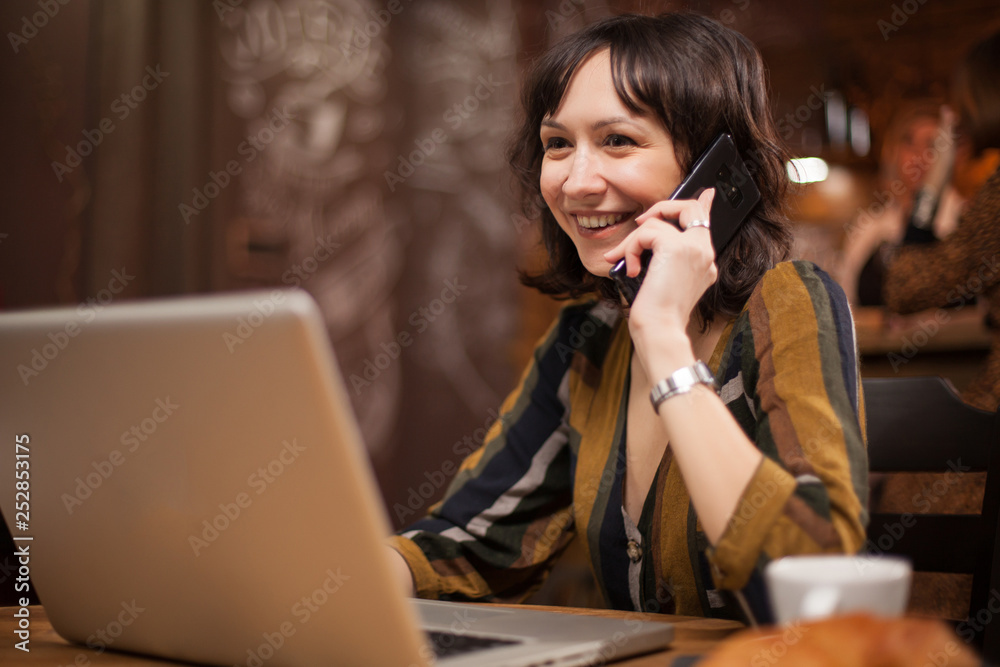 Pretty young woman smiling while talking on the phone and using her laptop in a coffee shop Pretty young woman smiling while talking on the phone and using her laptop in a coffee shop