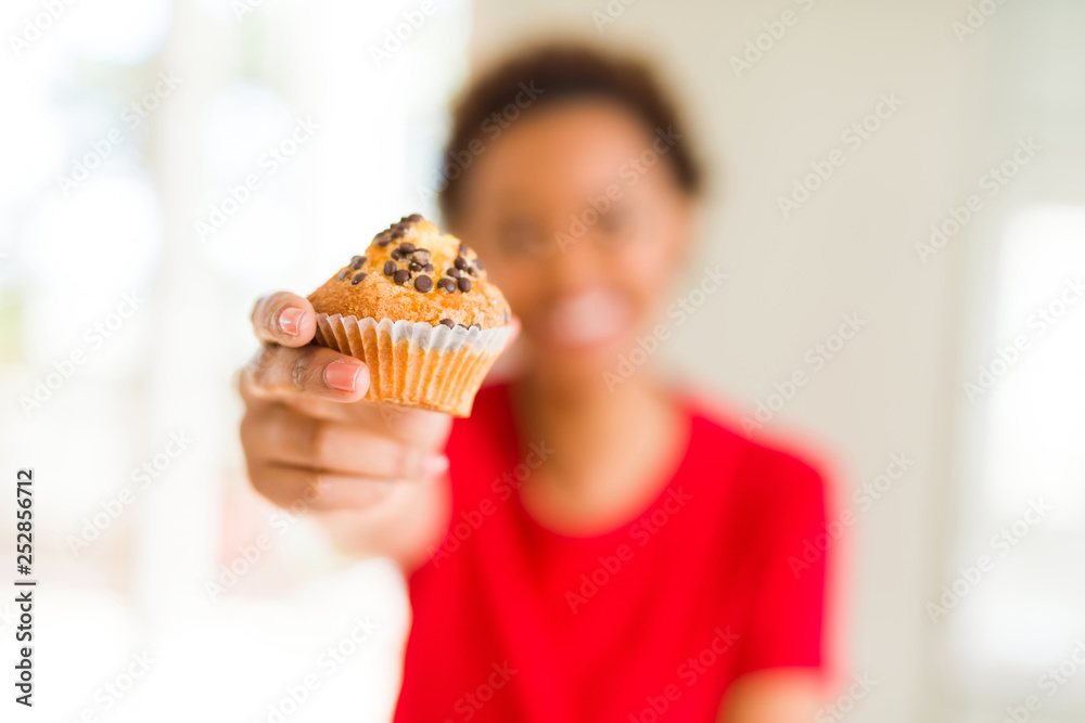 Young african american woman eating chocolate chips muffins with a happy face standing and smiling with a confident smile showing teeth Young african american woman eating chocolate chips muffins with a happy face standing and smiling with a confident smile showing teeth