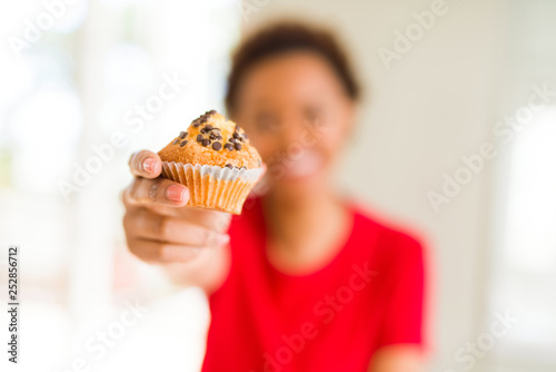 Young african american woman eating chocolate chips muffins with a happy face standing and smiling with a confident smile showing teeth
