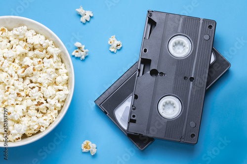Two black VHS tapes, bowl of popcorn and scattered popcorns on a blue background. Movie/ film industry. Retro/ vintage technology. Entertainment.