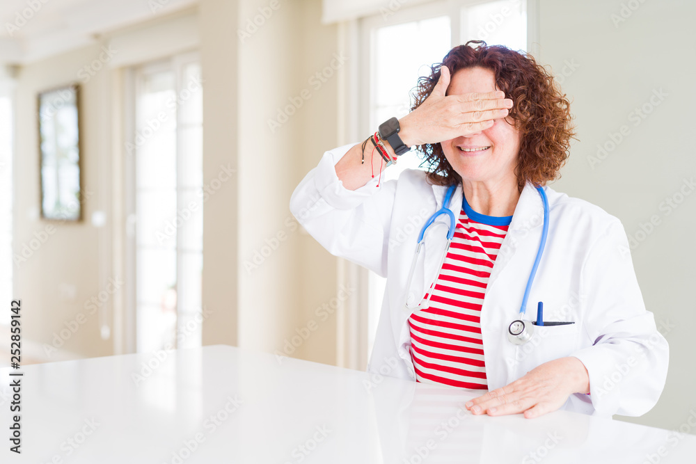Senior doctor woman wearing medical robe at the clinic smiling and laughing with hand on face covering eyes for surprise. Blind concept. Senior doctor woman wearing medical robe at the clinic smiling and laughing with hand on face covering eyes for surprise. Blind concept.