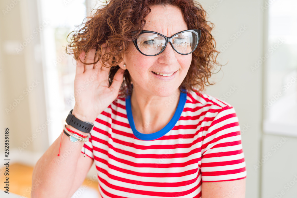 Beautiful senior woman wearing glasses and casual t-shirt smiling with hand over ear listening an hearing to rumor or gossip. Deafness concept. Beautiful senior woman wearing glasses and casual t-shirt smiling with hand over ear listening an hearing to rumor or gossip. Deafness concept.