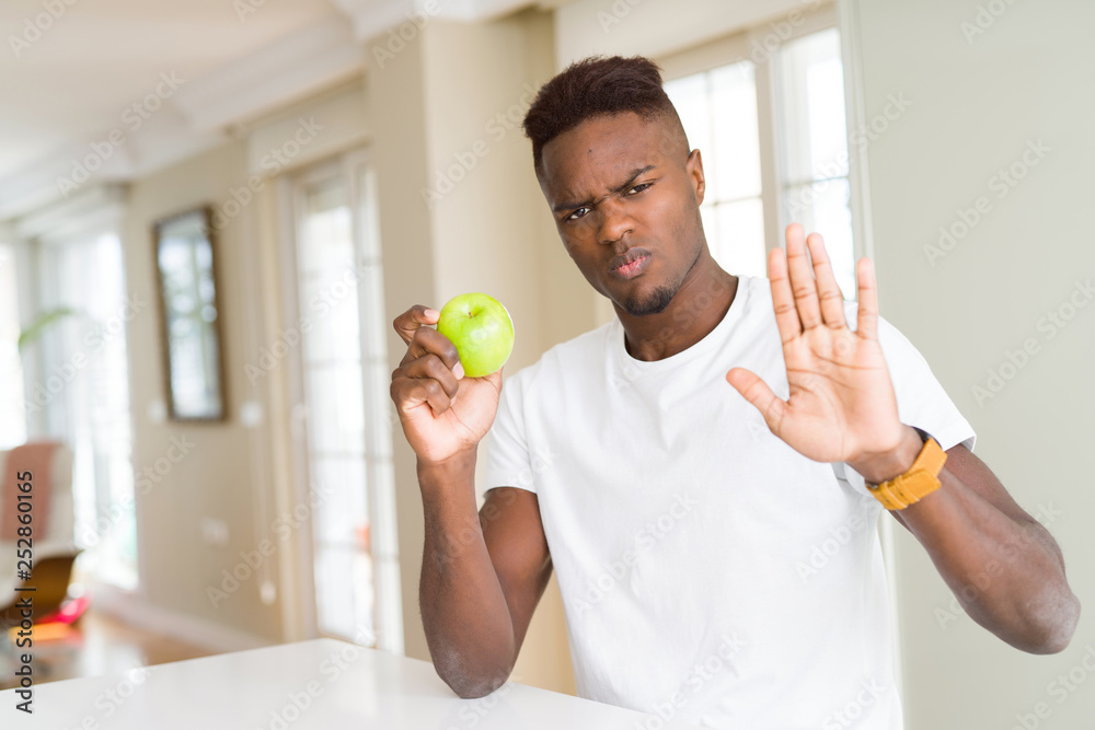 Young african american man eating fresh green apple with open hand doing stop sign with serious and confident expression, defense gesture Young african american man eating fresh green apple with open hand doing stop sign with serious and confident expression, defense gesture