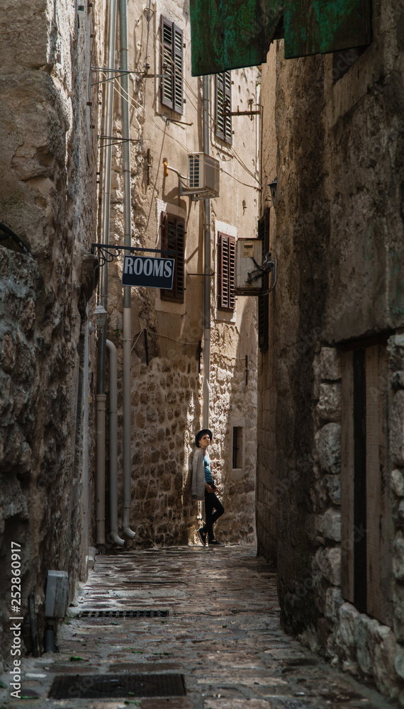 A girl stands in a narrow street in an old European city with a cobbled road and stone walls.