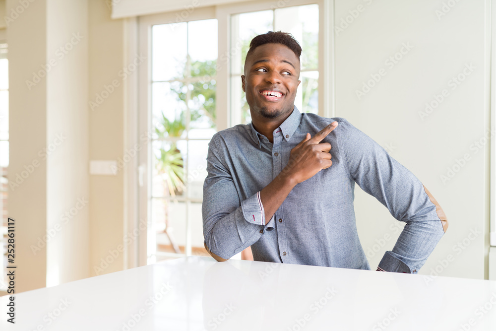 Handsome african american man on white table cheerful with a smile of face pointing with hand and finger up to the side with happy and natural expression on face Handsome african american man on white table cheerful with a smile of face pointing with hand and finger up to the side with happy and natural expression on face