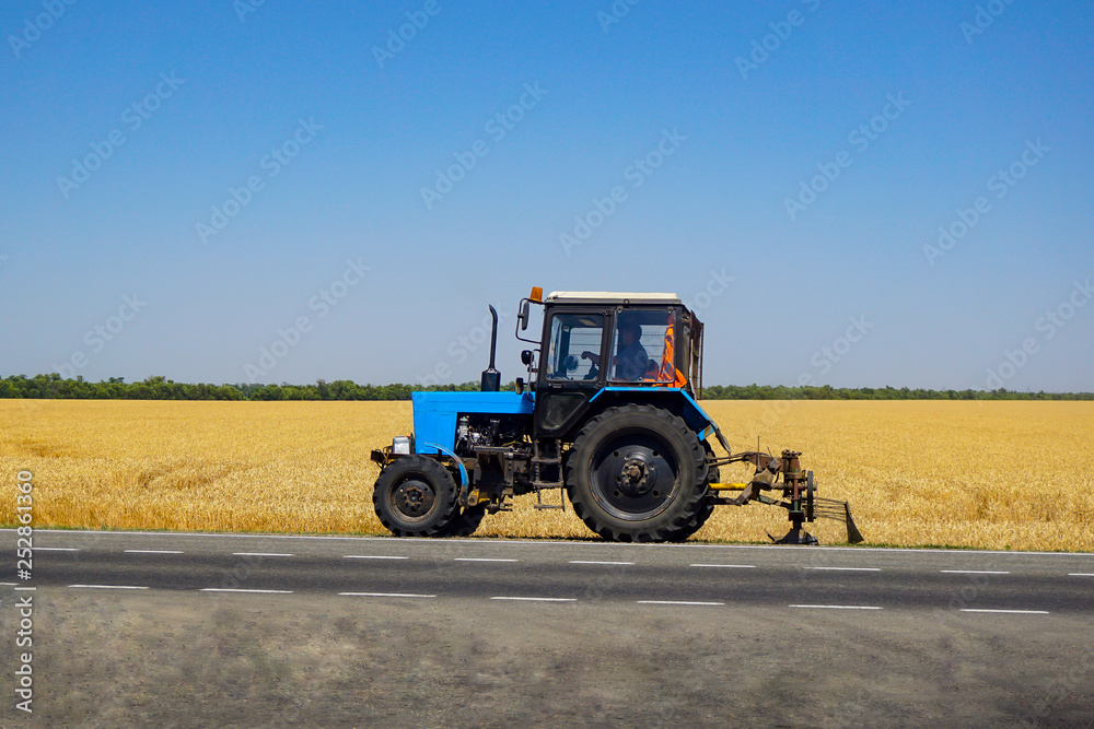 Fototapeta premium Ciągnik kosi trawę w pobliżu pola. Rolnik zasadził ogródek warzywny. Niekończące się pola Rosji w regionie Rostów na tle autostrady.
