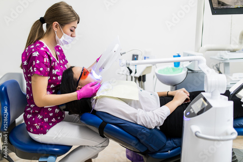 Close-up portrait of female patient at woman dentist in the clinic. Teeth whitening procedure with ultraviolet light UV lamp.