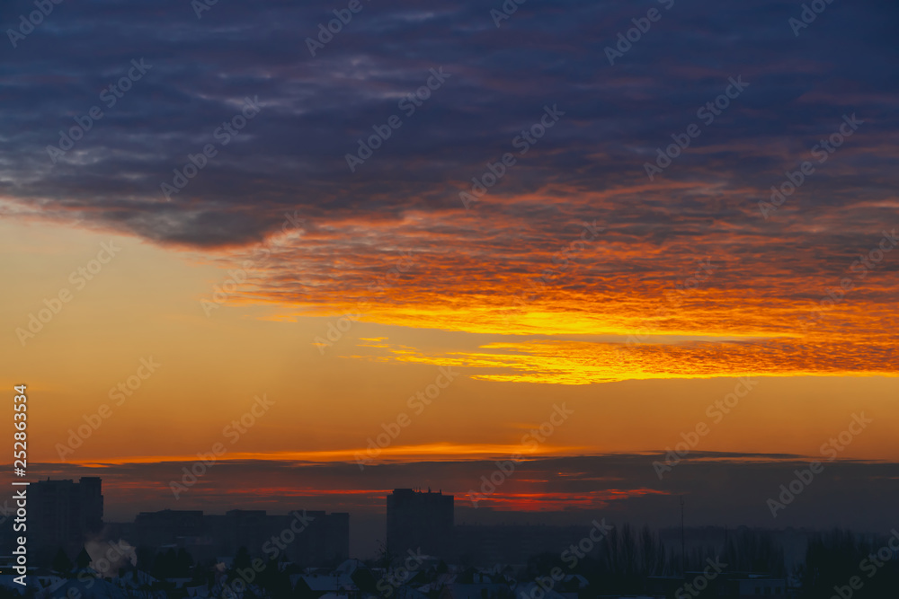 Cityscape with wonderful varicolored fiery dawn. Amazing dramatic multicolored cloudy sky. Dark silhouettes of city building roofs. Atmospheric background of sunrise in overcast weather. Copy space.