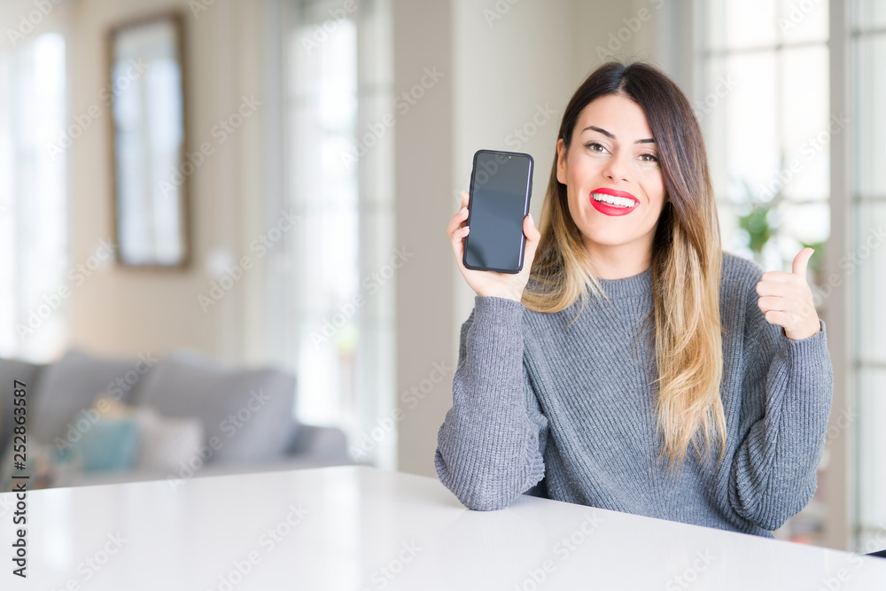 Young beautiful woman showing smartphone screen at home pointing and showing with thumb up to the side with happy face smiling