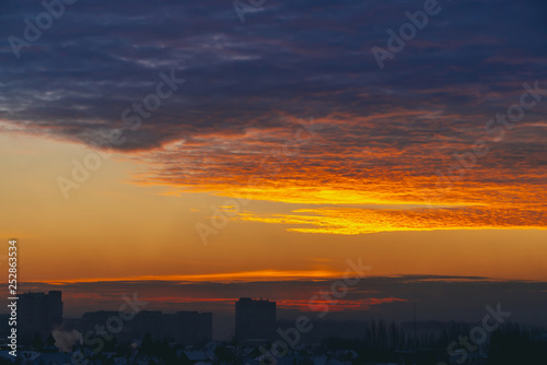 Cityscape with wonderful varicolored fiery dawn. Amazing dramatic multicolored cloudy sky. Dark silhouettes of city building roofs. Atmospheric background of sunrise in overcast weather. Copy space.