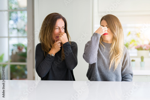 Beautiful family of mother and daughter together at home smelling something stinky and disgusting, intolerable smell, holding breath with fingers on nose. Bad smells concept.