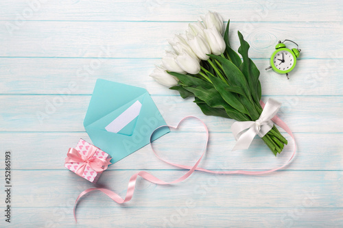 A bouquet of white tulips and a pink ribbon in the form of a heart with a gift box  love note and color envelope on blue wooden boards