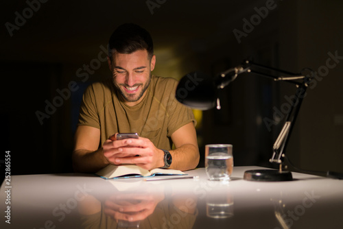 Young handsome man studying at home   using smartphone  looking at the phone smiling